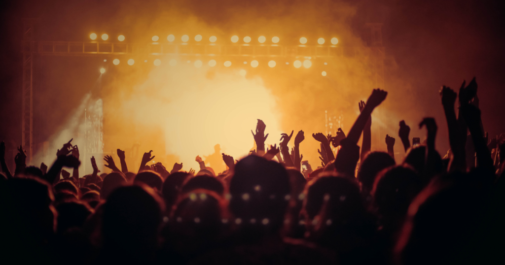Imagine cu o mulțime de oameni la un concert. Publicul ridică mâinile spre scenă, iar luminile portocalii și fumul creează o atmosferă intensă și energică.