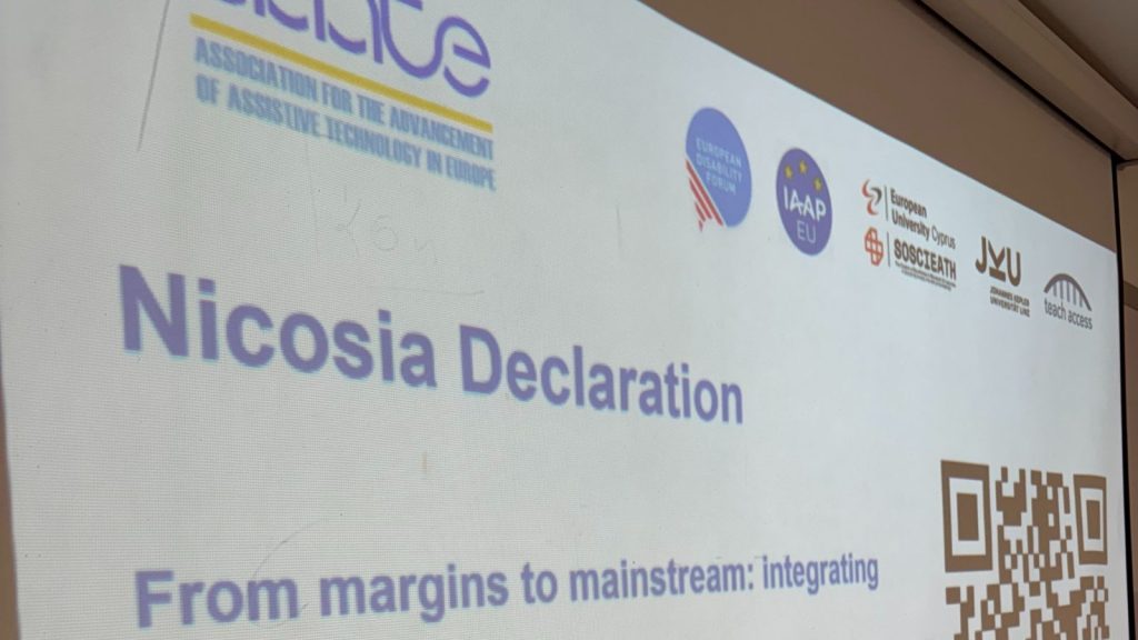Fotografie proiectată pe un ecran, cu titlul «Nicosia Declaration». Subtitlu: «From margins to mainstream: integrating». În partea stângă sus apare logo-ul AAATE – Association for the Advancement of Assistive Technology in Europe, iar în dreapta sunt mai multe logo-uri de organizații partenere: European Disability Forum, IAAP EU, European University Cyprus, SOSCETATH, Johannes Kepler Universität Linz și Teach Access. În partea dreaptă jos este afișat un cod QR.