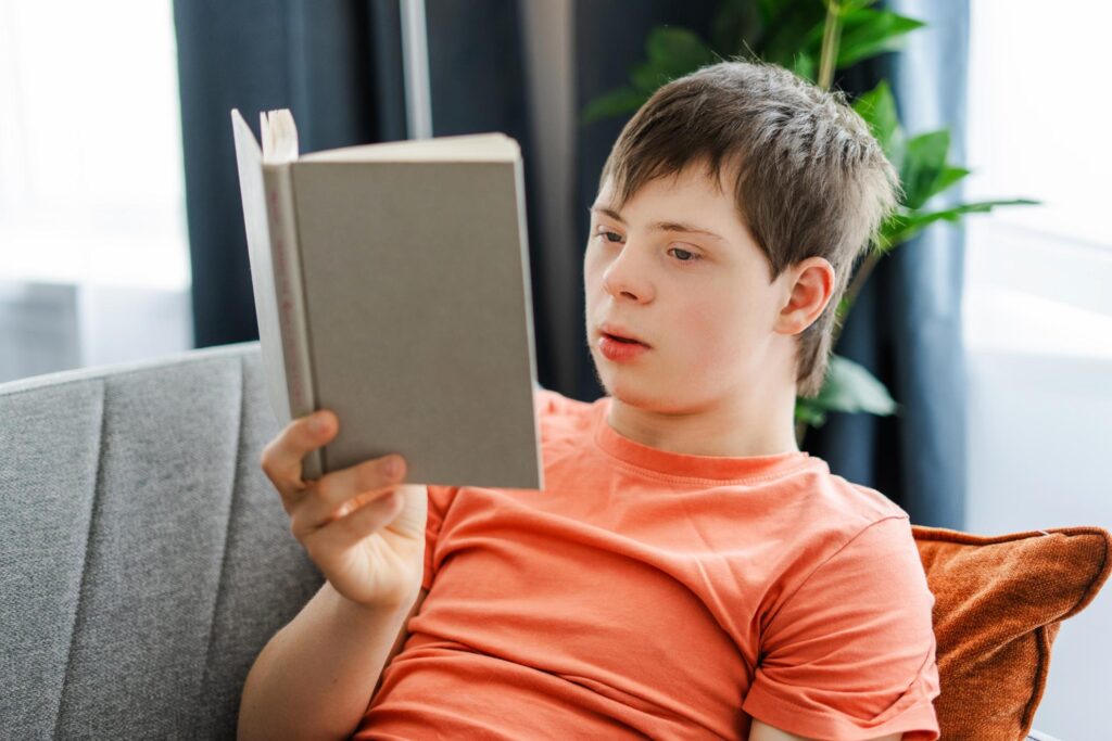 Un adolescent stă pe o canapea și citește atent o carte cu copertă gri, într-un living luminos, având în fundal o plantă verde și perdele închise la culoare.