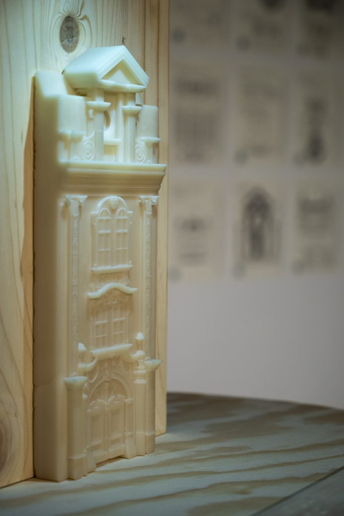 În această imagine se regăsește o placă de lemn pe care stă o fațadă de clădire în formă 3D de culoare crem