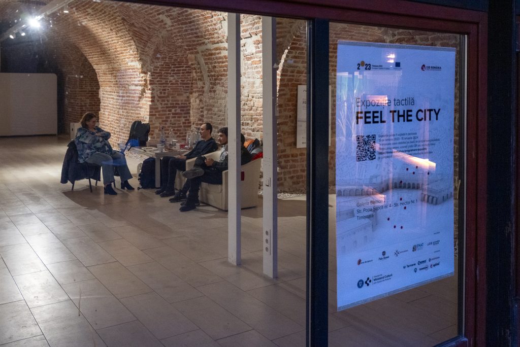 Imaginea reprezintă intrarea în clădire, în plan principal fiind geamyl cu afișul Feel the City, iar în plan secundar îndepărtat sunt câteva persoane care stau pe canapele