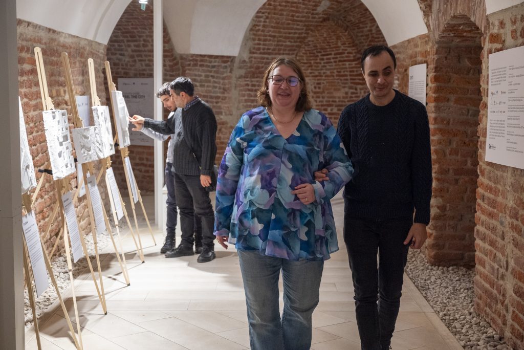 În imagine sunt 4 persoane, 3 bărbați și o femeie. În plan secundar, doi bărbați ating machetele 3D din expoziția înșirată pe șevalet, în plan principal femeia îl conduce pe bărbatul nevăzător care o ține de braț