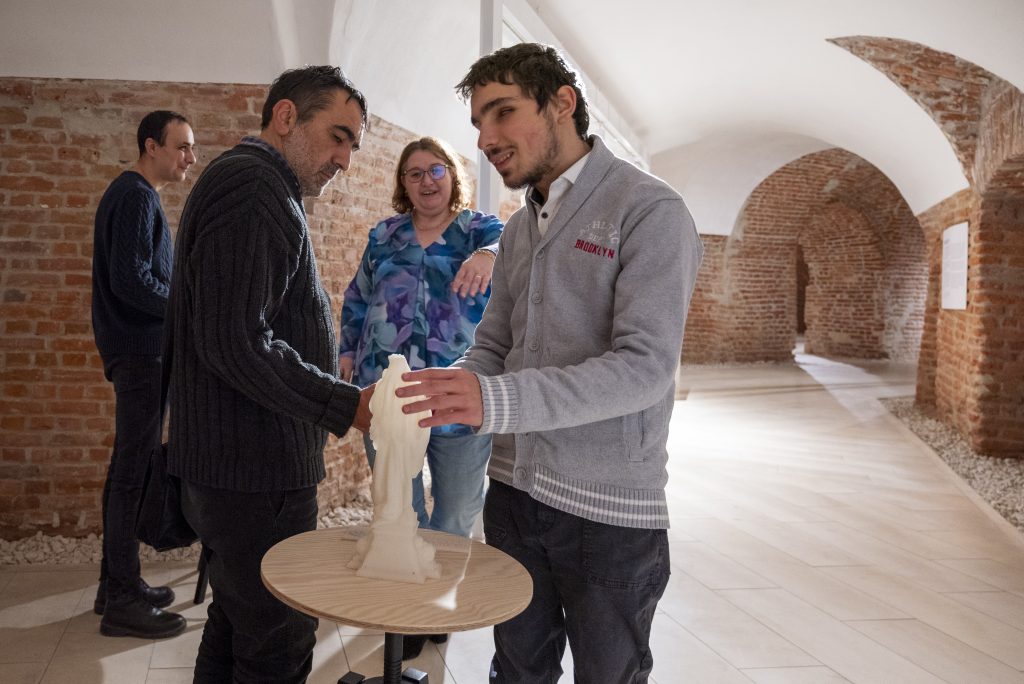 În imagine sunt 4 persoane, trei bărbați și o femeie, două în prim plan, celelalte două în planul din spate. Cele din față sunt doi bărbați nevăzători care atinge o machetă 3D care reprezintă pe Maica Domnului