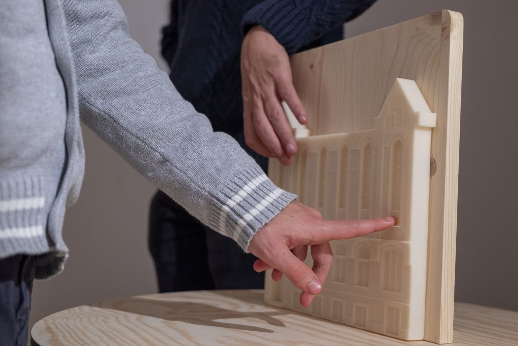 În imagine sunt două brațe a două persoane diferite care examinează tactil o fațadă de clădire în format 3D pe o placă de lemn