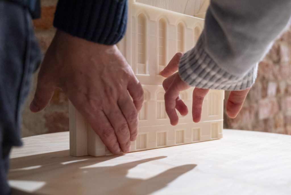 În imagine sunt două brațe a două persoane diferite care examinează tactil o fațadă de clădire în format 3D pe o placă de lemn
