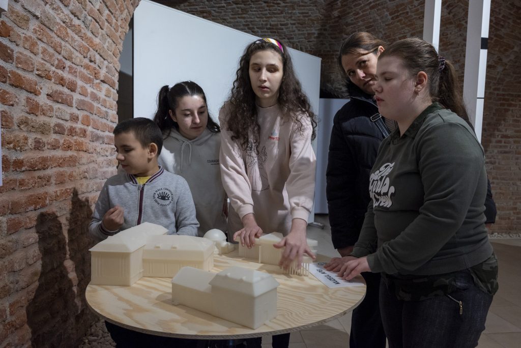 În imagine se află 5 persoane, 4 fete și un băiețel, unii dintre ei fiind nevăzători. Copiii ating machetele 3D de pe măsuță pentru a simți formele în relief