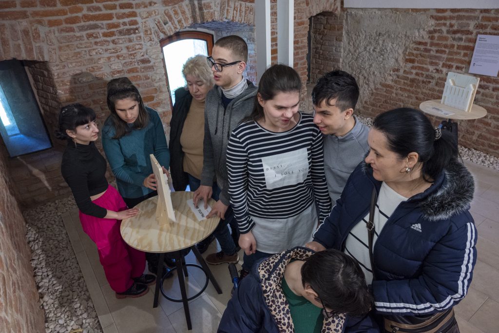 În imagine se află 8 persoane, copii nevăzători, fete și băieți împreună cu îndrumătorii acestora, copiii atingând macheta 3D pentru a simți formele în relief