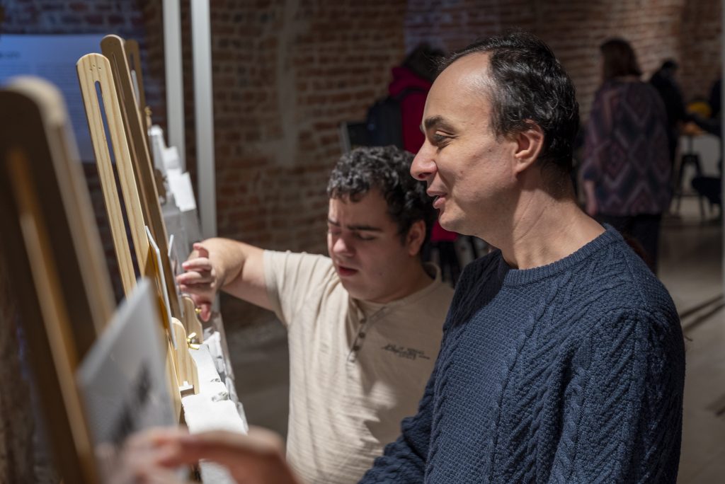 În această imagine sunt doi bărbați, unul este nevăzător și se bucură de experiența tactilă a machetelor 3D din expoziție