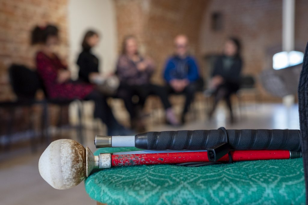 În această imagine se află un bărbat și o femeie care stau pe scaune, imaginea fiind blur, iar în prim planul imaginii cu focus se află pe o masă un baston de culoare roșu și negru