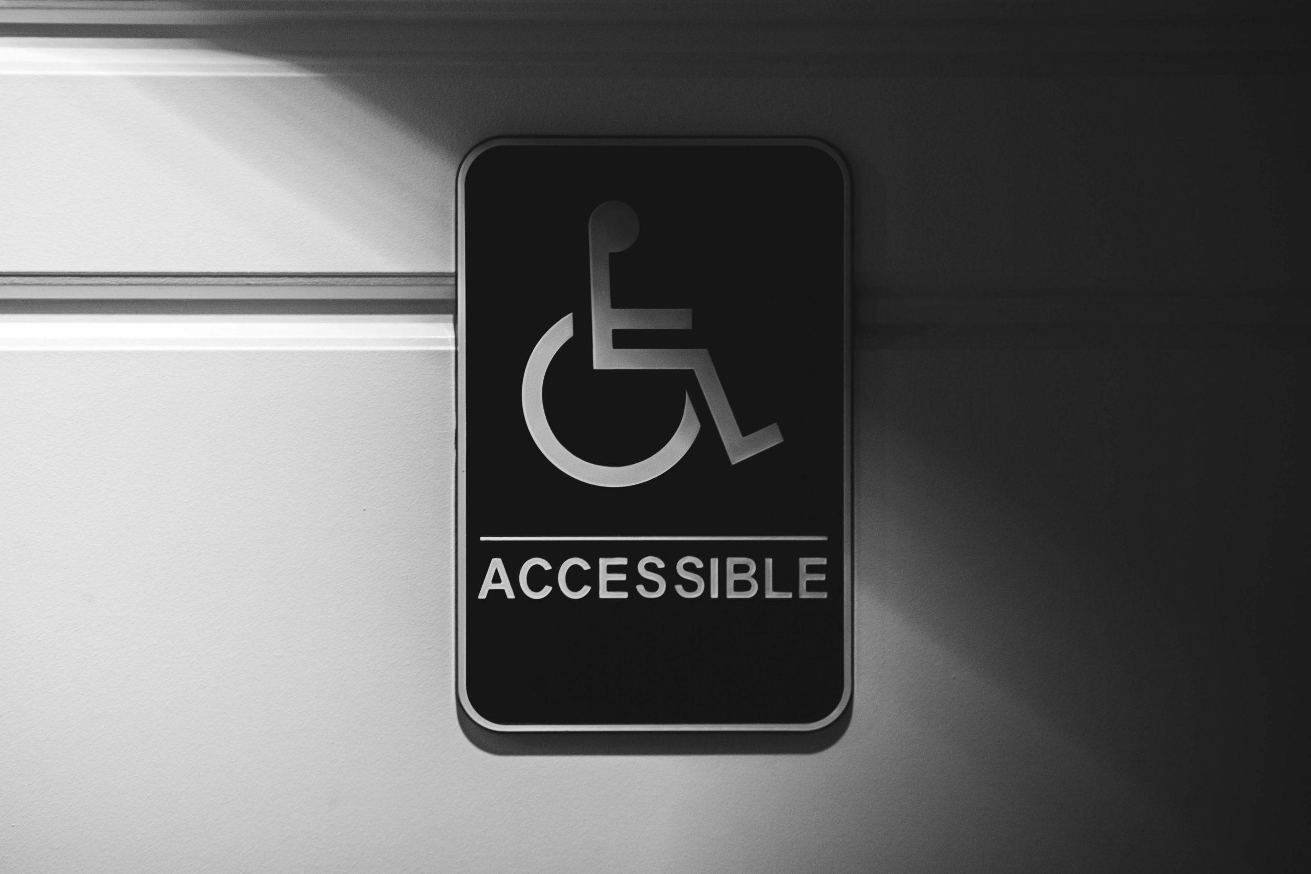imaginea are un perete alb și pe acest perete este un panou negru cu semnul dizabilitatii pe el unde scrie in limba engleza accesibil