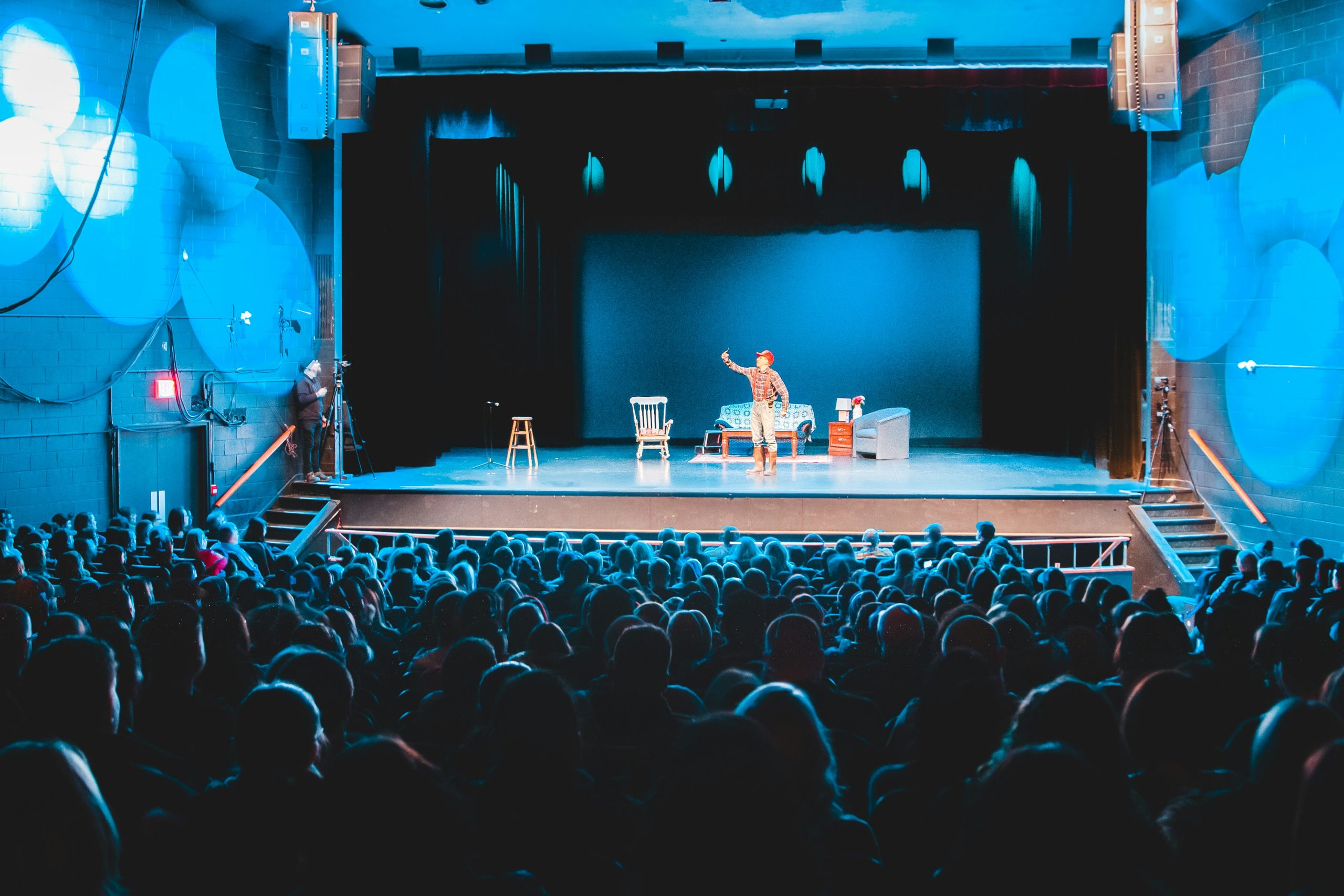 imaginea ilustrează o încăpere de teatru cu mai mulți oameni care vizionează o piesă de teatru, pe scenă fiind un bărbat care interpretează un personaj