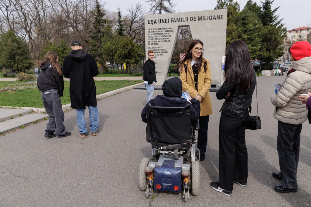 Un grup de persoane discută în fața unei instalații urbane cu mesajul „Ușa unei familii de milioane”. O persoană în scaun rulant stă în centrul imaginii, iar altele sunt în jurul ei, în picioare, vorbind și ținând pliante. În fundal se văd copaci și o alee dintr-un parc.