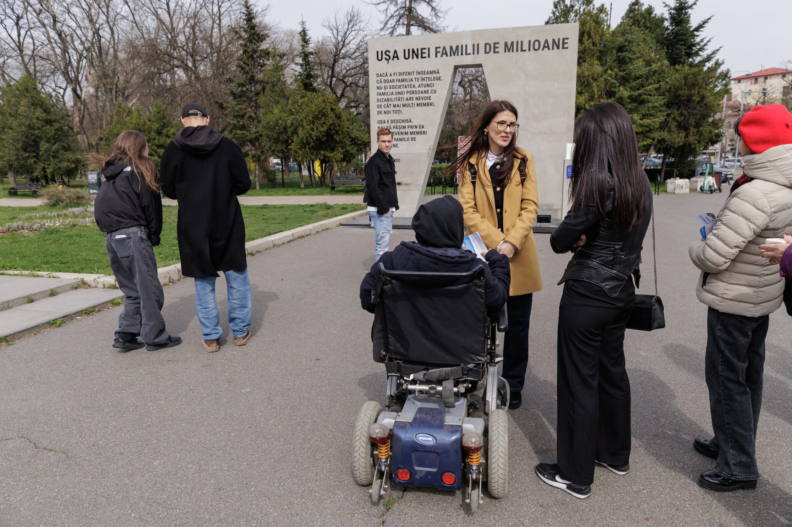 Un grup de persoane discută în fața unei instalații urbane cu mesajul „Ușa unei familii de milioane”. O persoană în scaun rulant stă în centrul imaginii, iar altele sunt în jurul ei, în picioare, vorbind și ținând pliante. În fundal se văd copaci și o alee dintr-un parc.