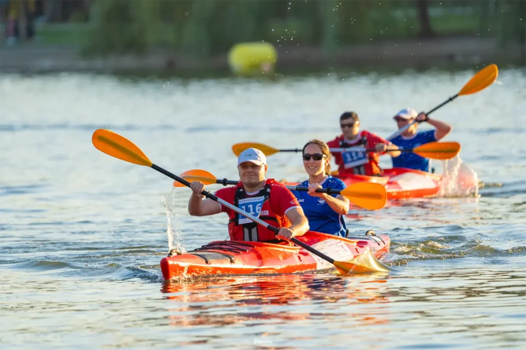 Două caiace duble cu patru sportivi vâslind pe un lac, purtând veste de salvare și folosind padele portocalii, în timpul unei activități sportive pe apă, într-un cadru natural.